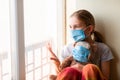 Sad little girl with teddy bear toy both in protective masks sitting on the window at home . Coronavirus quarantine Royalty Free Stock Photo