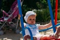 Sad, little girl in a white dress and hat, riding on a swing, summer sun and heat. playground. childhood, serenity. negative Royalty Free Stock Photo