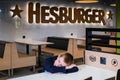 Sad and lonely boy is sitting at the table while his parents have gone to buy food. Logo of Finnish company Hesburger on the wall