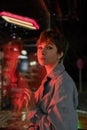 Sad lonely woman in red neon light leaning on shop window on street at night, looking at camera