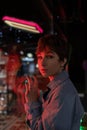 Sad lonely woman in red neon light leaning on shop window on street at night, looking at camera