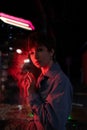 Sad lonely woman in red neon light leaning on shop window on street at night, looking at camera