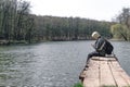 A sad man is sitting alone on the pier by the lake. Forest in the background. hood on his head. backpack. in hands hold phone. Royalty Free Stock Photo