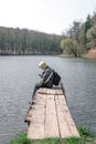 A sad man is sitting alone on the pier by the lake. Forest in the background. hood on his head. backpack. in hands hold phone. Royalty Free Stock Photo