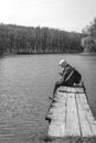 A sad man is sitting alone on pier by the lake. Forest black and white. hood on his head. backpack. in hands hold phone. Emotions Royalty Free Stock Photo