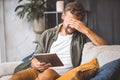 Sad man sitting on sofa home, holding tablet PC, making facepalm gesture. Frustration and disappointment on face