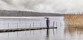 Man standing under umbrella on wooden pier on autumn rainy gray day over of backdrop of lake and forest. Royalty Free Stock Photo