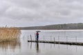 Man standing under umbrella on wooden pier on autumn rainy gray day over of backdrop of lake and forest. Royalty Free Stock Photo