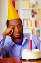 Sad man wearing blue shirt and hat sitting by table with cake in front, single candle burning, looking bored depressed Royalty Free Stock Photo