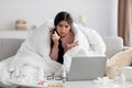 Sad millennial indian female wrapped in blanket with napkin, is ill at home in living room interior, looks in laptop