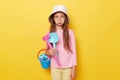 Sad offended little cute girl in panama holding beach sandbox toys rake and shovel isolated over yellow background looking at Royalty Free Stock Photo