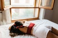 A sad and pensive young woman with ponytail, red top and white short lying in the bed looking out the window at home