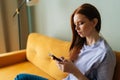 Sad red-haired young woman looking at smartphone screen expression face thinking about problem sitting on yellow couch Royalty Free Stock Photo