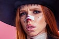 Sad redhaired ginger woman witch black hat with crazy surgery make up on pretty face in studio pink background.stop to