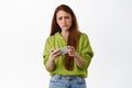 Sad redhead girl losing video game on mobile phone, watching upsetting video on smartphone and grimacing, frowning