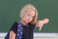 Sad schoolgirl with a backpack is showing a thumbs down in a classroom near green chalkboard. The child does not like the school. Royalty Free Stock Photo