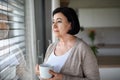 Sad senior woman standing and looking out through window indoors at home. Royalty Free Stock Photo