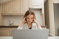 Sad student girl studying at home. Unhappy girl sitting at table in kitchen in apartment with laptop and looking at screen with Royalty Free Stock Photo