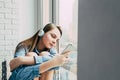 Sad teenager girl in blue shirt and modern headphones listens music and chatting with friends leaning on the window clutching her Royalty Free Stock Photo