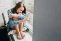 A sad teenager girl in a blue shirt sits near the window on the pillow and looks at the smartphone and chat with friends, hugging Royalty Free Stock Photo