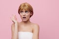 a sad, thoughtful woman stands on a pink background wrapped in a white towel and makes a negative emotional face