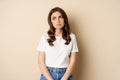 Sad and tired brunette woman looking drained and unamused, standing with pokerface against beige background