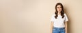 Sad and tired brunette woman looking drained and unamused, standing with pokerface against beige background