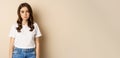 Sad and tired brunette woman looking drained and unamused, standing with pokerface against beige background