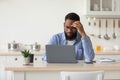 Sad tired pensive young black bearded guy in glasses works at laptop, ponders problem solution in scandinavian kitchen Royalty Free Stock Photo