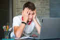 Sad tired teenager in white shirt sitting behind desk in kitchen  near laptop and study. Depressed Boy in earphones makes homework Royalty Free Stock Photo