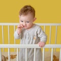 Sad toddler baby boy in the crib gnawing a nail, yellow studio background. A tired child in pajamas