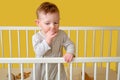 Sad toddler baby boy in the crib gnawing a nail, yellow studio background. A tired child in pajamas