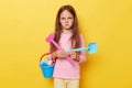 Sad unhappy bored little cute girl with dark hair holding beach sandbox toys rake and shovel isolated over yellow background Royalty Free Stock Photo