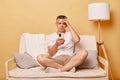 Sad unhappy brunette adult man wearing casual clothing sitting on sofa against beige wall using smartphone reading bad news Royalty Free Stock Photo