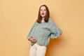 Sad unhealthy young pregnant woman wearing knitted sweater standing isolated over beige background holding her tummy feeling pain Royalty Free Stock Photo