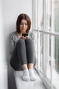 Sad upset caucasian millennial female sitting on windowsill at home and reading message on phone with bad news Royalty Free Stock Photo