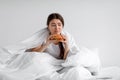 Sad upset depressed european millennial woman eating burger in bed under duvet at home alone Royalty Free Stock Photo