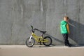 Sad upset little boy standing near concrete wall with his bicycle on sunny day. Urban lifestyle