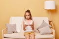 Sad upset little girl wearing casual attire sitting on sofa against beige wall using cell phone loosing game level looking at Royalty Free Stock Photo