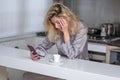 Sad woman drink coffee and using phone at home in morning. Tired female madel chatting smartphone in sexy pajama at