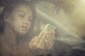 Sad woman looking in the mobile phone and reading message in the window with rain drop in the car. Royalty Free Stock Photo