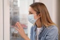 Sad woman in protective mask looking out of window indoors. Staying at home during coronavirus pandemic Royalty Free Stock Photo