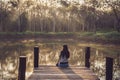 A sad woman sits floating on an old wooden bridge that juts out into a pond in nature. Lonely sad woman