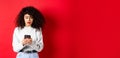 Sad and worried woman reading message on smartphone, receive bad news on social media, standing against red background Royalty Free Stock Photo