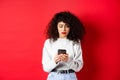 Sad and worried woman reading message on smartphone, receive bad news on social media, standing against red background Royalty Free Stock Photo