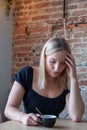 Sad young adult blonde woman sitting at window and holding mug in hands. Looking at her coffee in a cafe. Thinking about Royalty Free Stock Photo