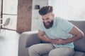 Sad young bearded man is suffering from stomach pain, ache is very hard. He is alone at home, sitting on couch Royalty Free Stock Photo