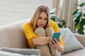 Sad young blonde european woman looks at phone, hugs her knees on sofa in living room interior Royalty Free Stock Photo