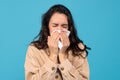 Sad young caucasian woman brunette suffers from runny, blows her nose at napkin, on blue background