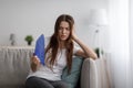 Sad young european woman suffer from hot and headache blowing fan at herself sitting on sofa in living room Royalty Free Stock Photo